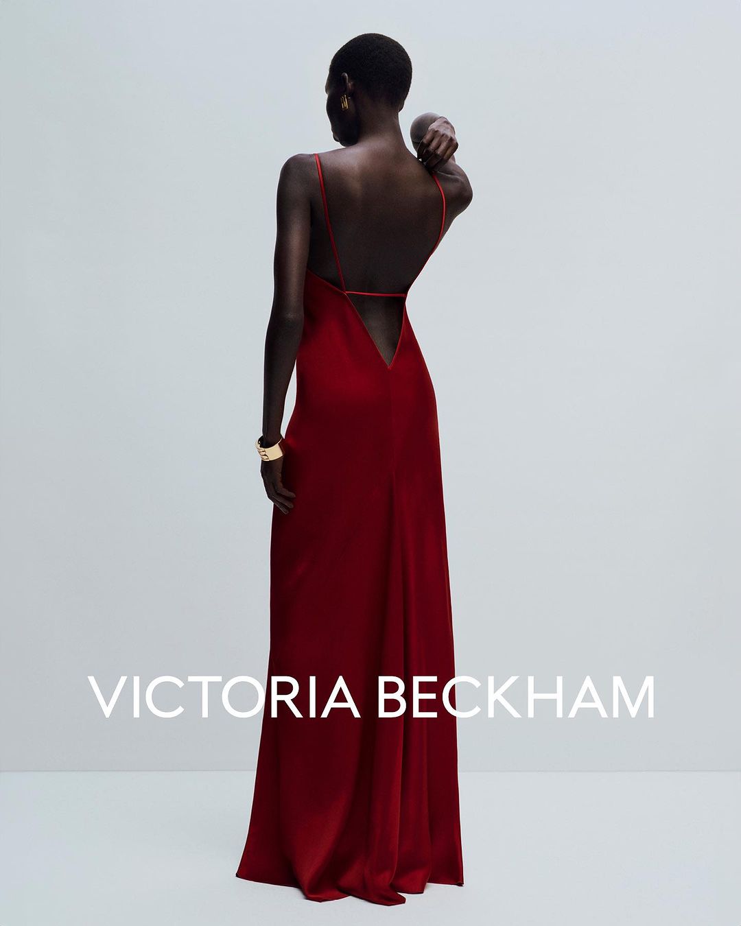 Back view of a black female model in a long, burgundy, sleeveless gown with thin straps and a V-shaped back, posing indoors.