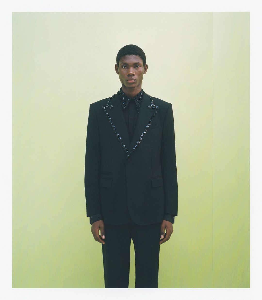 Fashionable Black man poses in a black suit with decorative trim against a pale yellow seamless backdrop in a full studio shot.