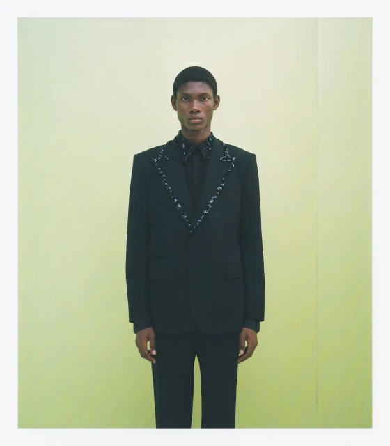 Fashionable Black man poses in a black suit with decorative trim against a pale yellow seamless backdrop in a full studio shot.