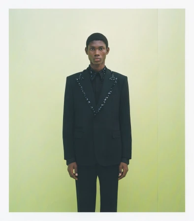 Fashionable Black man poses in a black suit with decorative trim against a pale yellow seamless backdrop in a full studio shot.