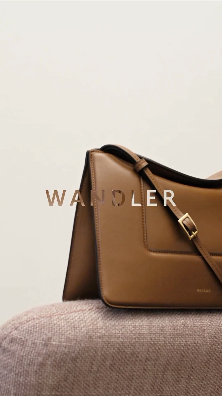 Tan leather shoulder bag with gold hardware and a branded logo on a mauve upholstered surface against a cream colored background.
