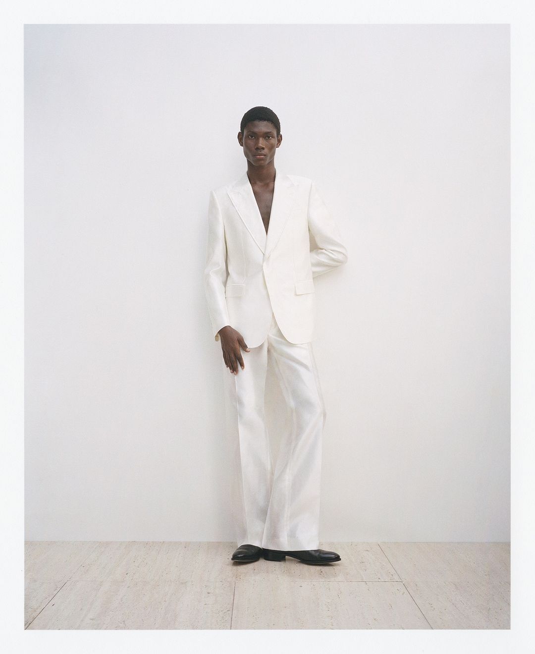A Black man in an open white suit stands in a photo studio wearing black shoes, with an elegant and modern aesthetic.