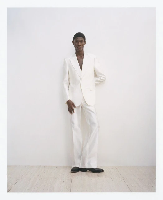 A Black man in an open white suit stands in a photo studio wearing black shoes, with an elegant and modern aesthetic.