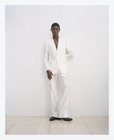 A Black man in an open white suit stands in a photo studio wearing black shoes, with an elegant and modern aesthetic.