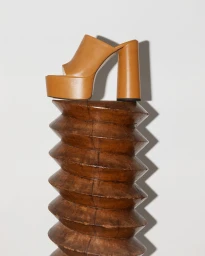 Tan leather platform mule resting on top of a carved wooden pedestal against a white background.