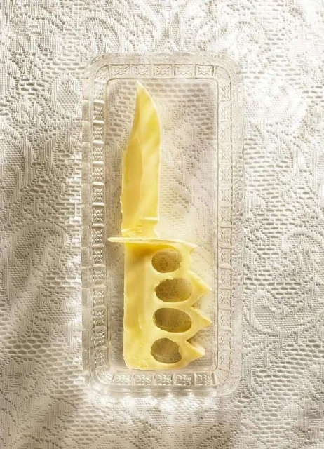 Overhead shot of a butter knife sculpted from butter inside a clear plastic tray against a patterned white background.