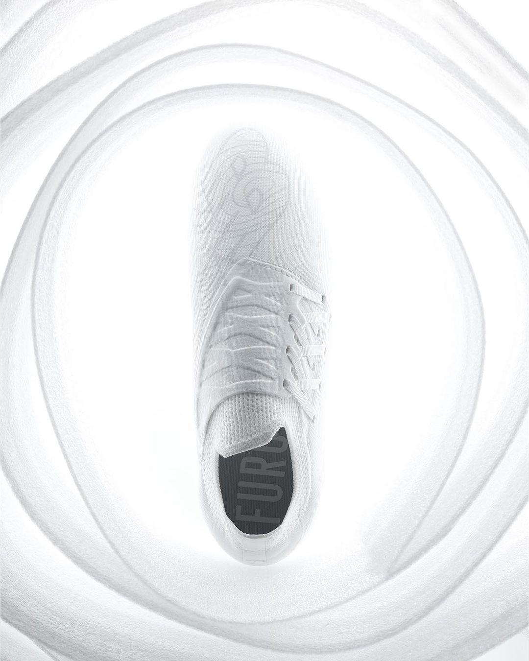 A single white sneaker is displayed overhead, surrounded by soft, out-of-focus fabric, creating a bright, minimalist footwear image.
