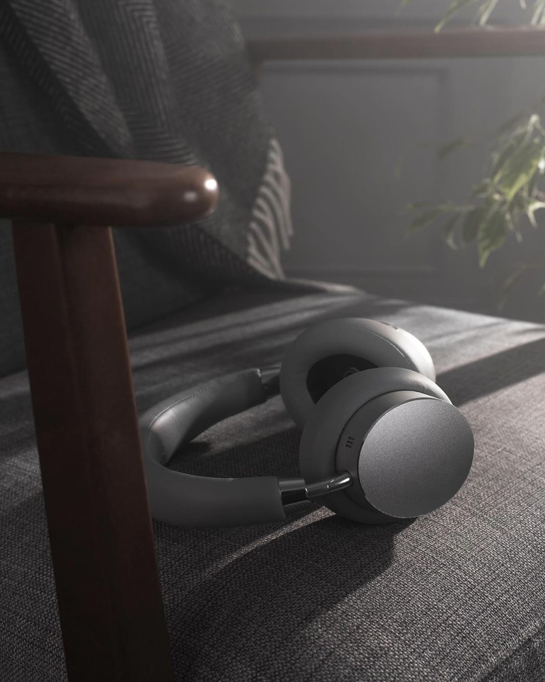Beige wireless headphones rest on a gray armchair, bathed in diffused daylight filtering through nearby indoor plants.
