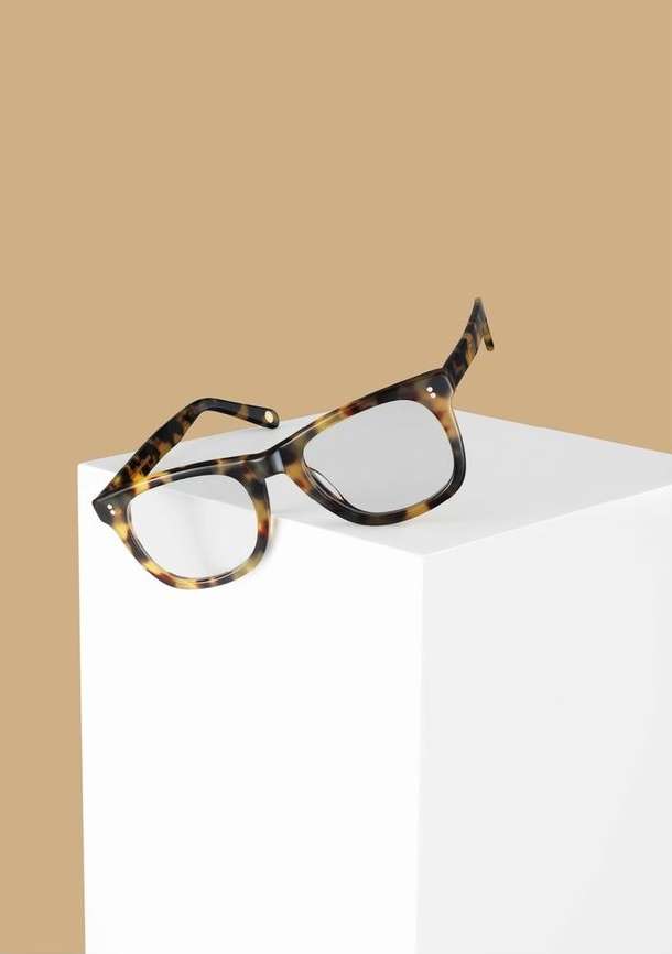 Studio shot of brown tortoise shell eyeglasses perched on a white cube against a tan background, minimalist product shot.