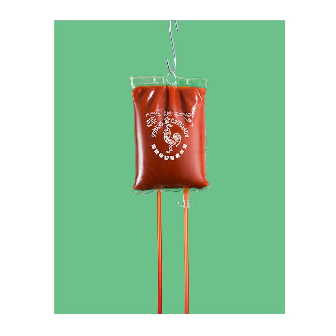 Sriracha sauce in a clear plastic bag hanging from a metal hook, resembling an IV drip, on a light green background.