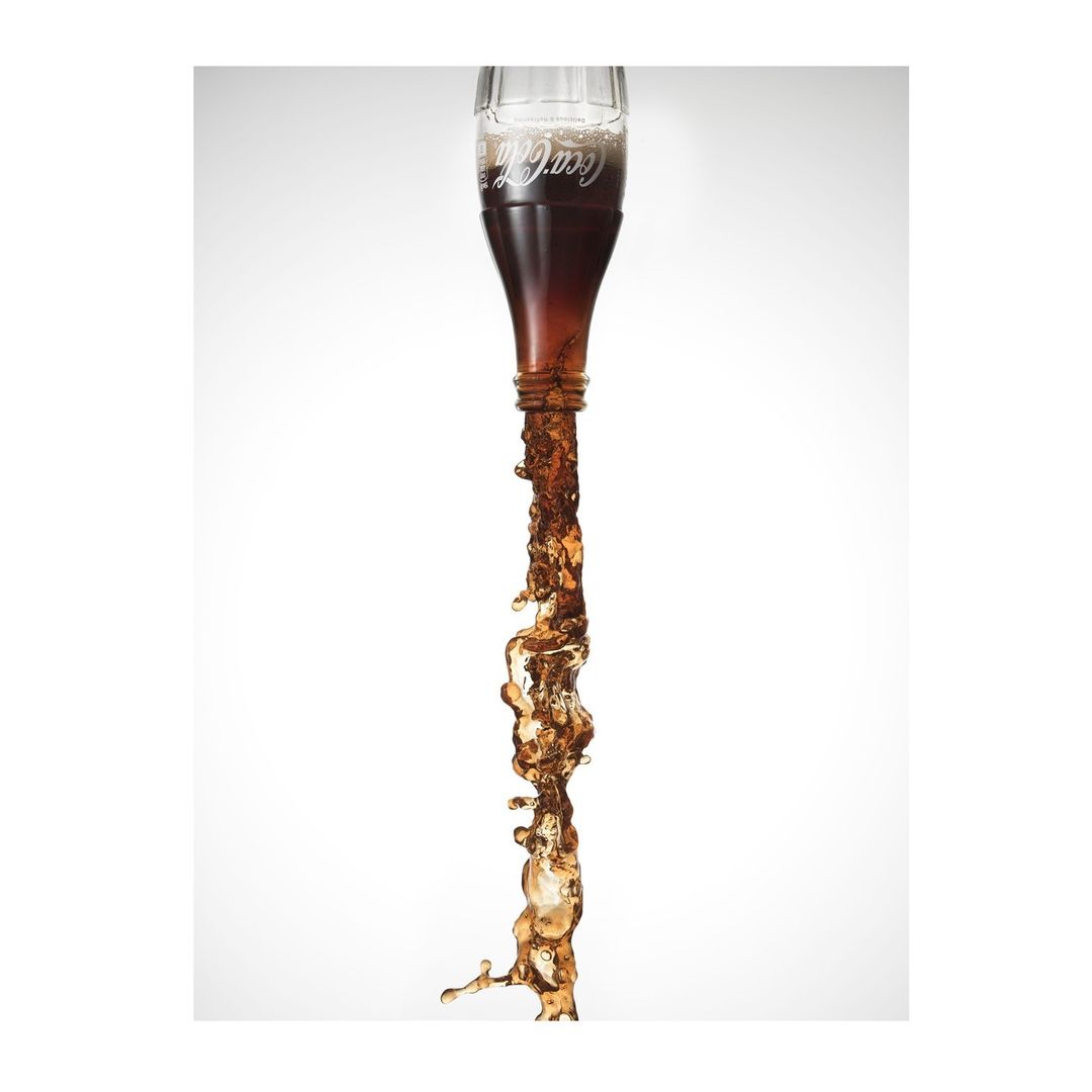 Coca-Cola bottle is inverted, and soda is captured mid-pour, creating a dynamic splash against a bright, neutral background.