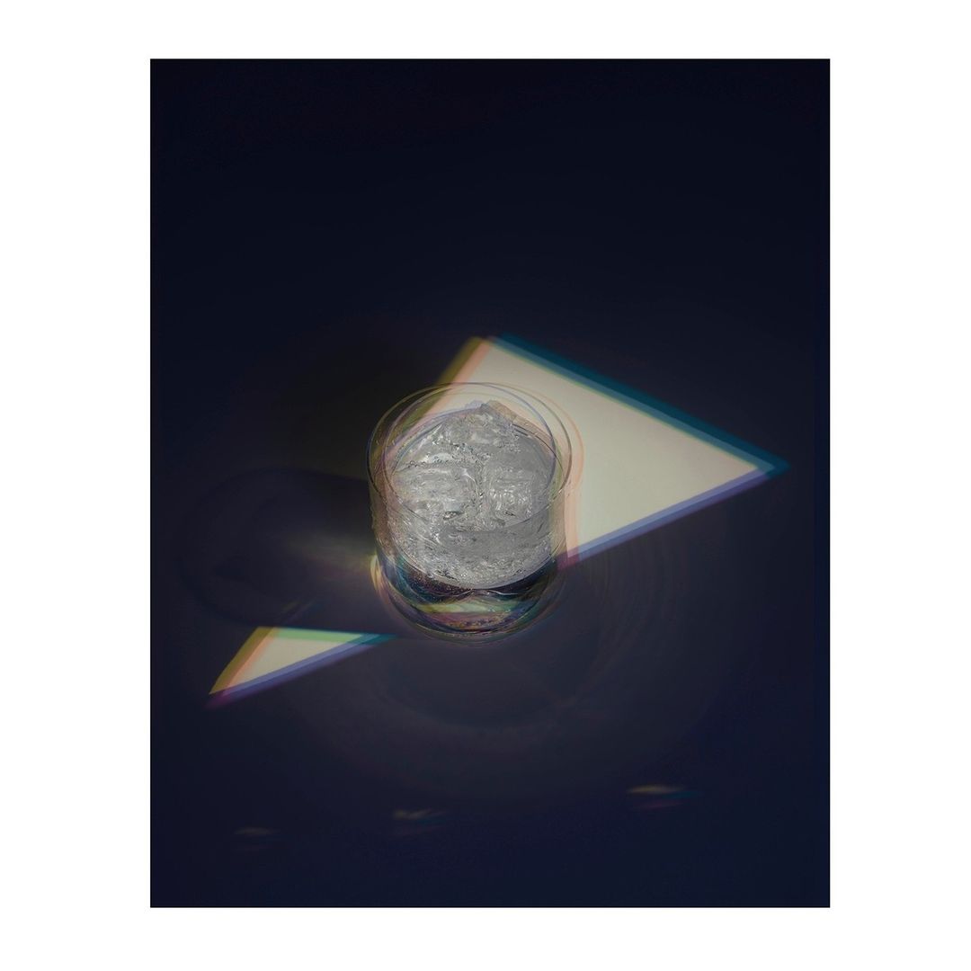 A clear glass with ice sits on a dark surface, light catching the ice and glass in an eye-level shot with interesting shadows.