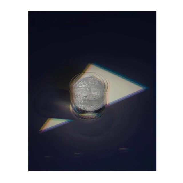 A clear glass with ice sits on a dark surface, light catching the ice and glass in an eye-level shot with interesting shadows.
