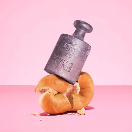 A still life photograph shows a 7kg weight crushing a bun against a pink background, creating a visually striking and potentially symbolic image.