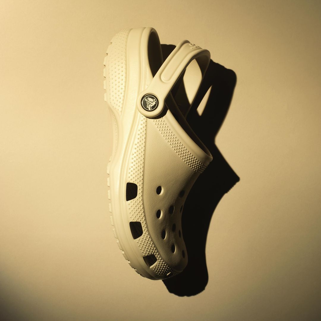 A beige Crocs shoe casts a dark shadow against a solid beige background, captured in a studio shot. The image is a product still life.