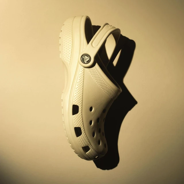 A beige Crocs shoe casts a dark shadow against a solid beige background, captured in a studio shot. The image is a product still life.