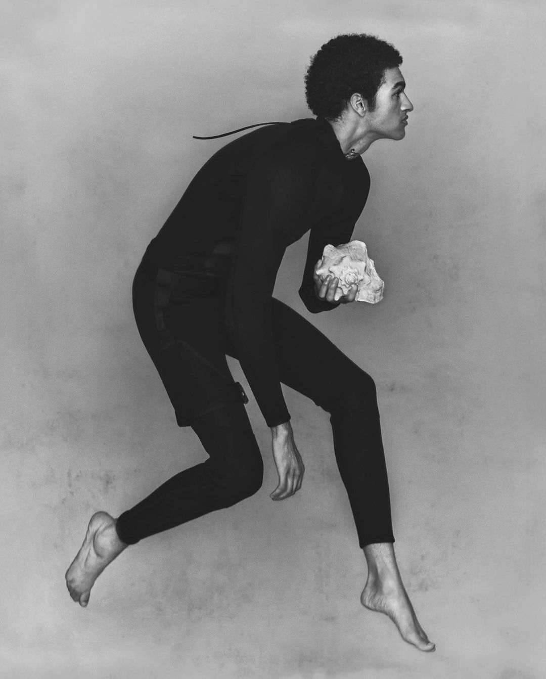 An adult man with an afro hairstyle in black athletic wear leaping in a monochrome studio shot, holding a light-colored rock in his hand.