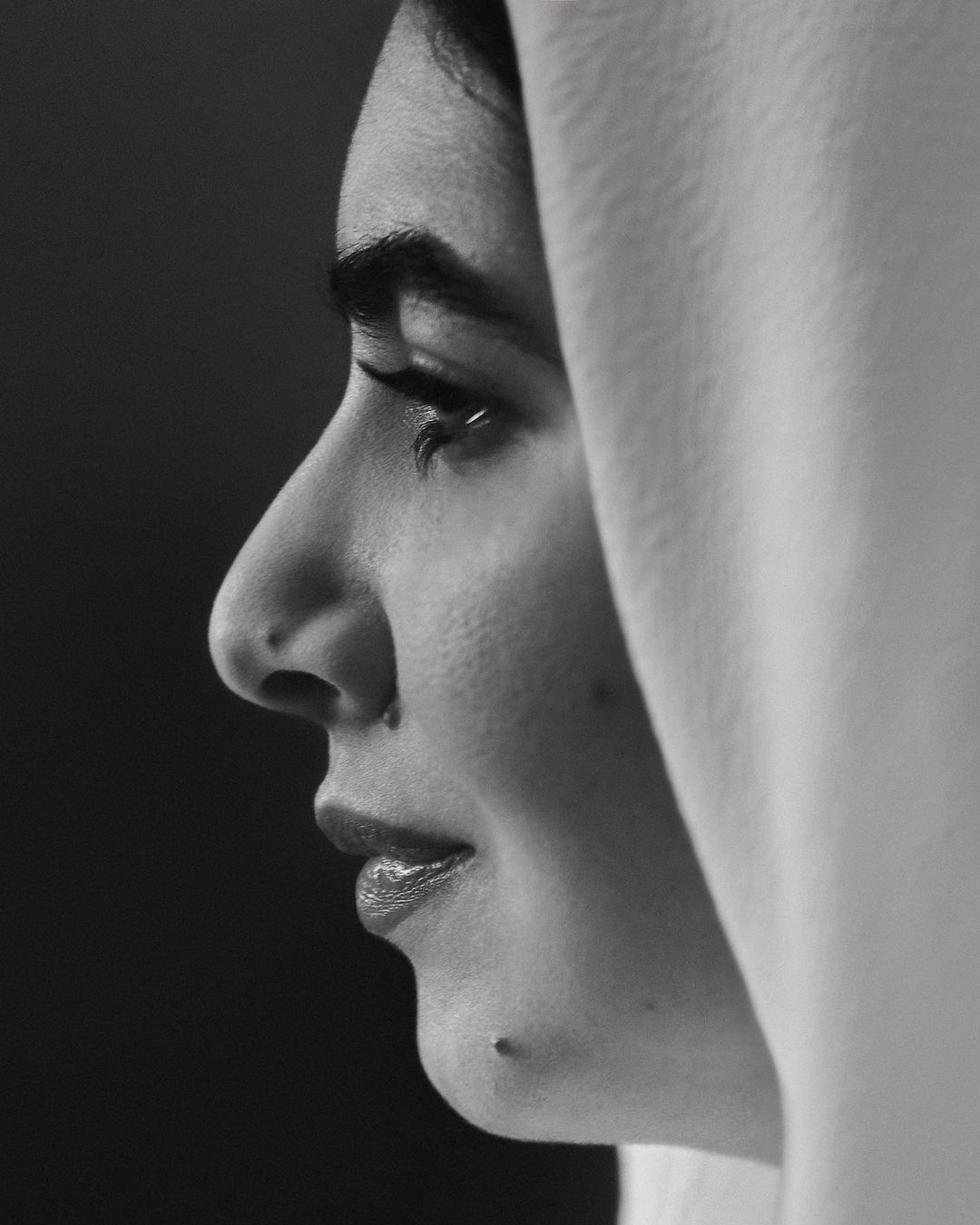 Close-up black and white portrait of a woman in profile wearing a light colored head covering against a dark background with soft lighting.