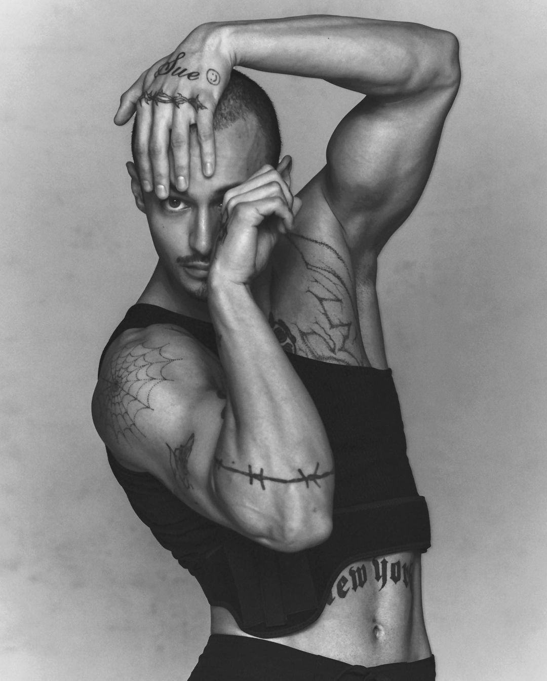 Black and white studio shot of muscular adult male posing with hand tattoos and a tank top in front of a gray backdrop.