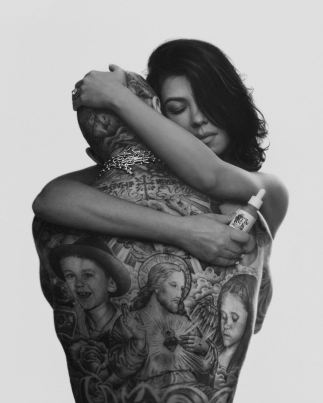Monochrome portrait of Kourtney Kardashian embracing a heavily tattooed man, set against a clean white backdrop with beauty product.