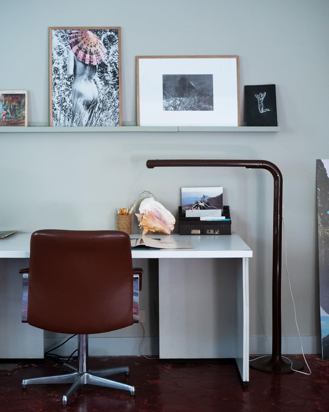 Interior shot features a brown leather office chair, white desk, shelving with framed pictures, and a lamp in a room with dark red flooring.