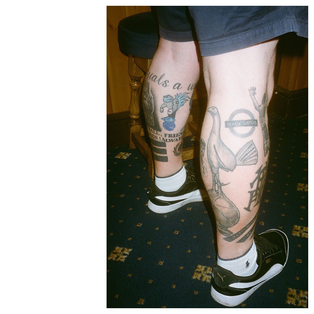 Tattooed legs of a person, wearing black sneakers with white soles, are positioned on a blue carpet with a gold pattern. The tattoos depict various images.