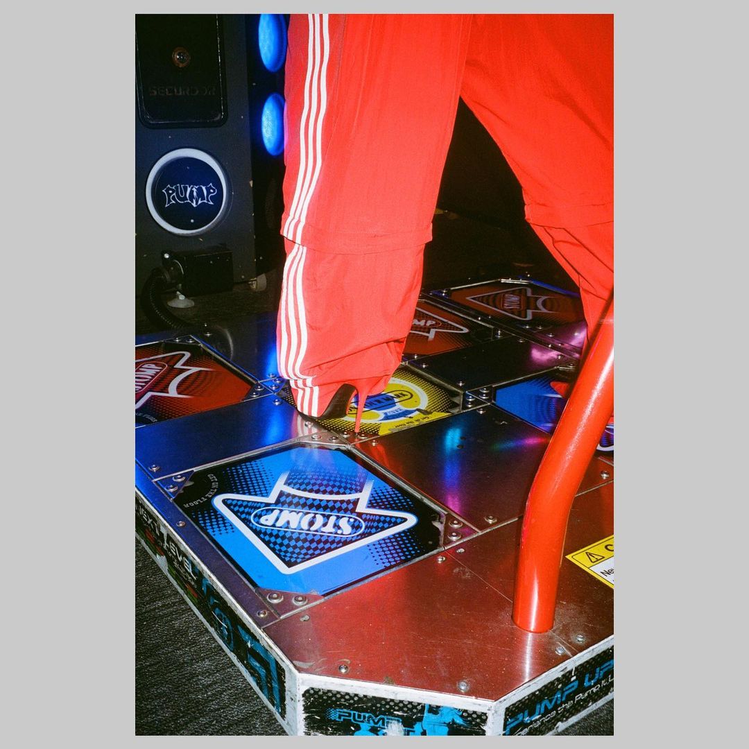 Person in red tracksuit pants plays dance game on lighted platform with directional arrows in an indoor arcade setting.