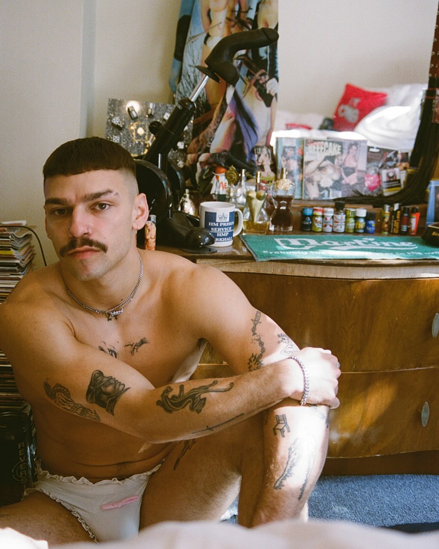 Shirtless man with tattoos and a mustache sits in underwear, indoors in ambient light with records, posters, and objects in background.