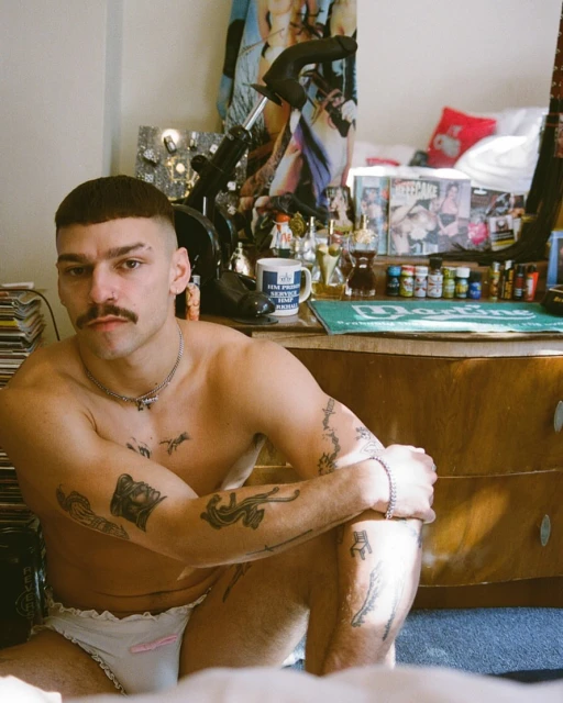 Shirtless man with tattoos and a mustache sits in underwear, indoors in ambient light with records, posters, and objects in background.