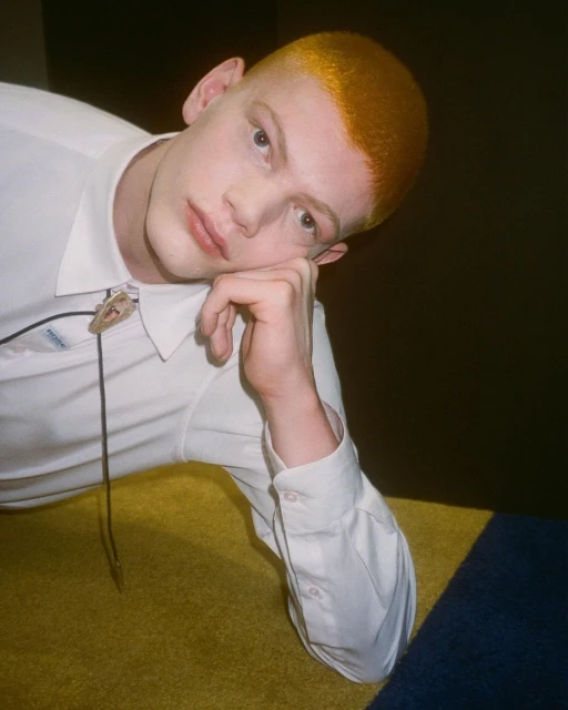 Close-up of a light-skinned adult male with yellow dyed short hair, wearing a white button-down shirt with a bolo tie, looking at the camera.