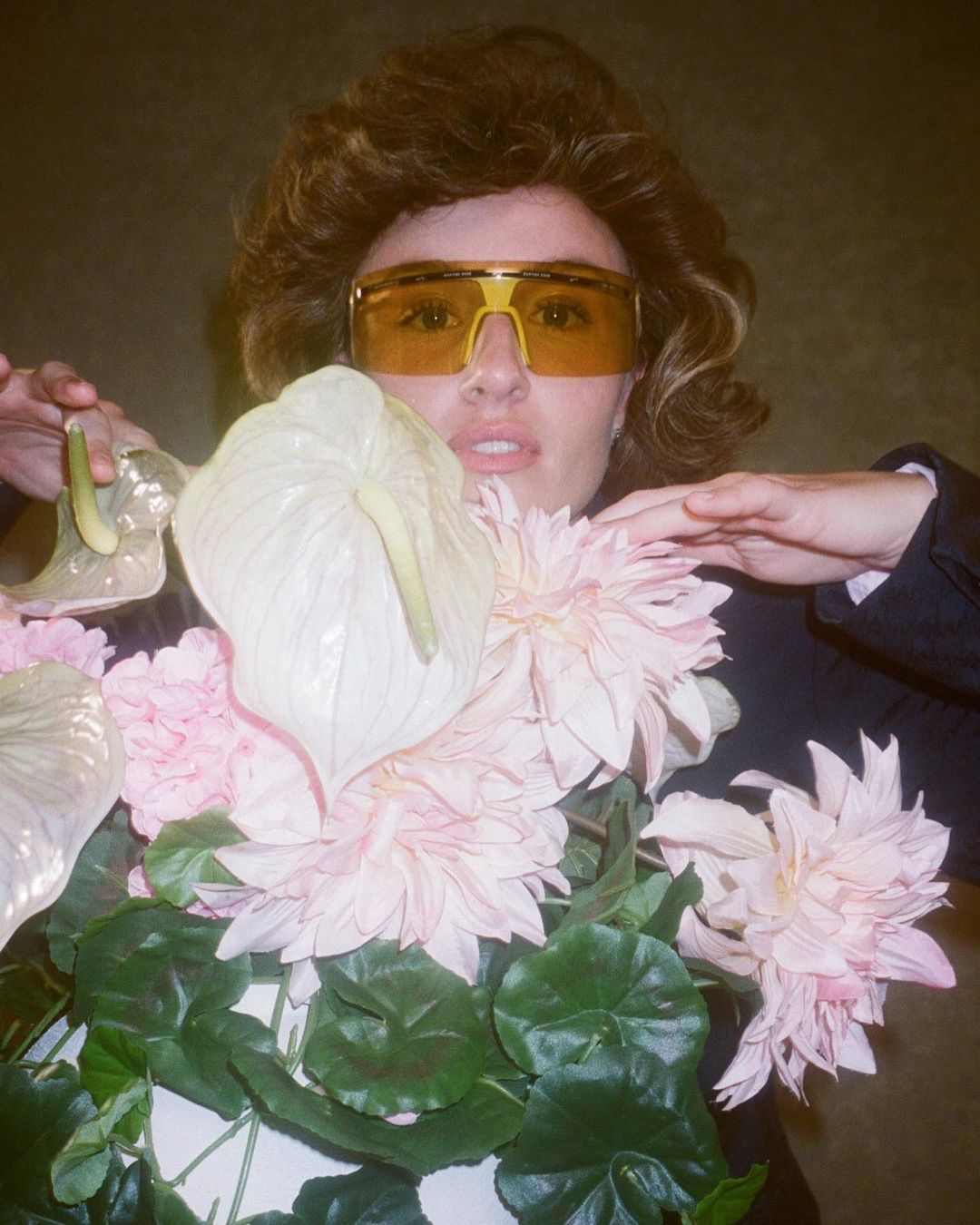 Woman in yellow sunglasses surrounded by flowers, posing in a studio. Fashion portrait with floral elements and a retro aesthetic.