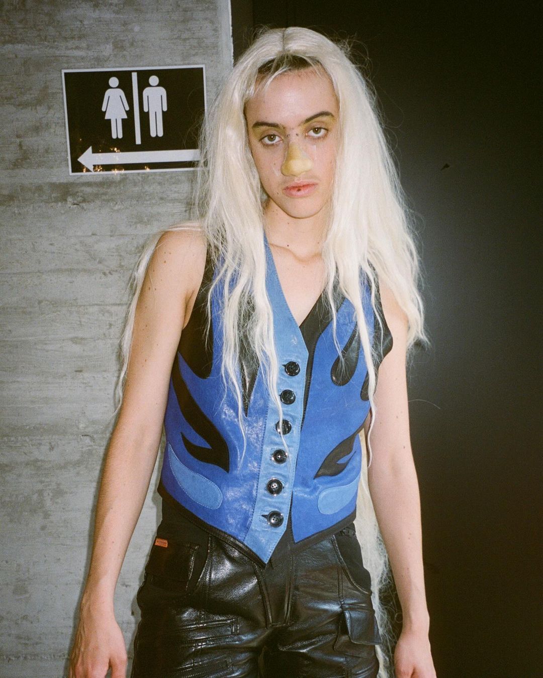 Non-binary person with pale blonde hair and a blue leather vest, photographed indoors with a restroom sign and black background.