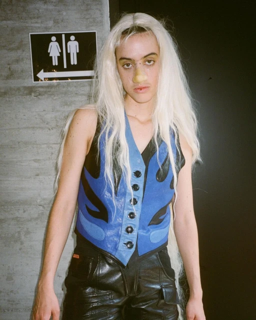 Non-binary person with pale blonde hair and a blue leather vest, photographed indoors with a restroom sign and black background.