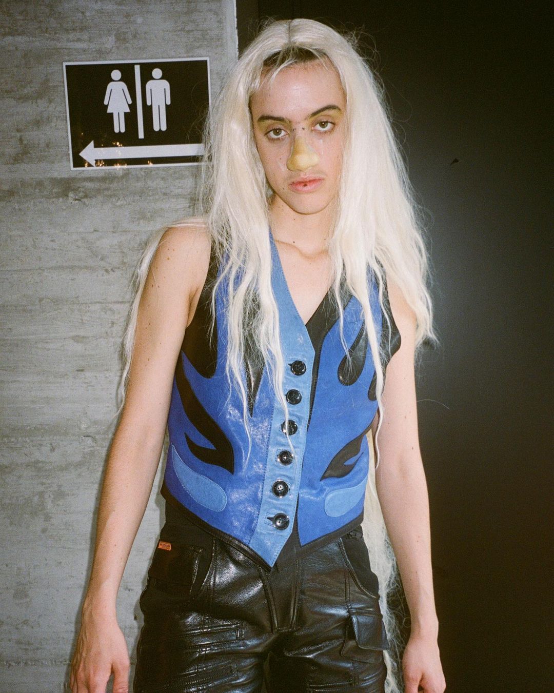 Person with long white wig, blue flame vest, and black leather shorts stands near a restroom sign in an indoor environment.