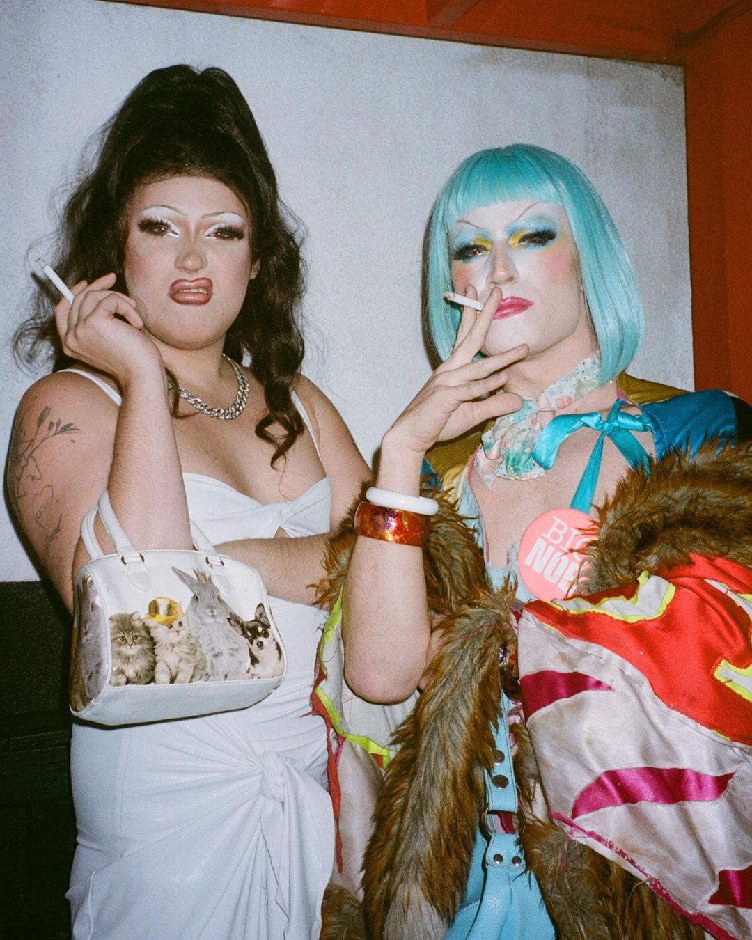 Two drag queens pose indoors, smoking cigarettes. One wears a white dress and holds a kitten purse, while the other wears teal wig and gown.