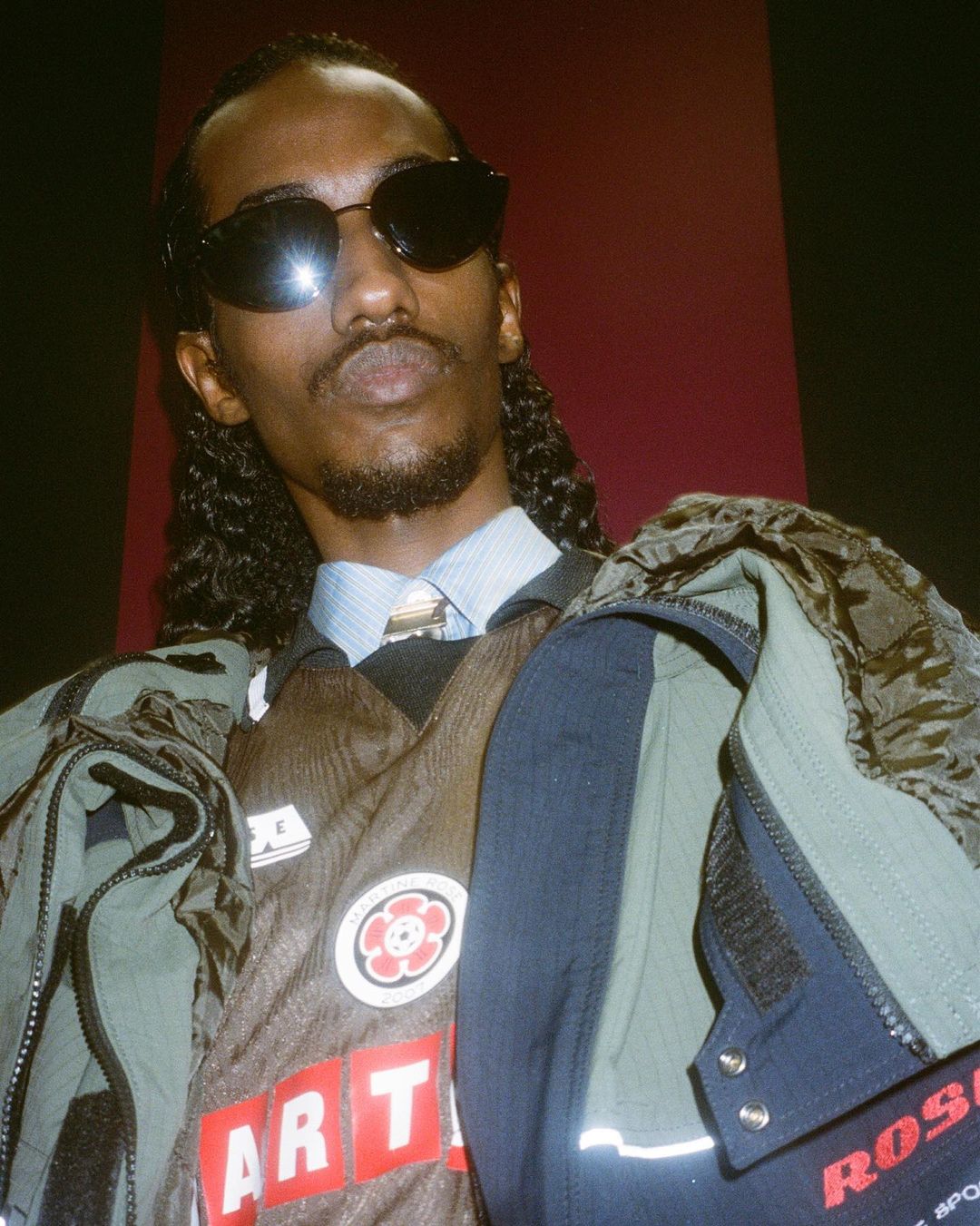 Man wearing sunglasses and layered jackets against a dark red wall photographed by Roxy Lee