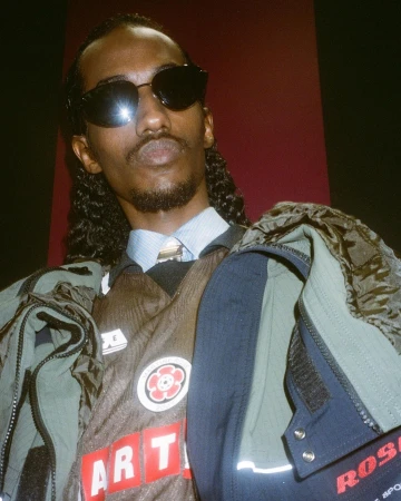Man wearing sunglasses and layered jackets against a dark red wall photographed by Roxy Lee