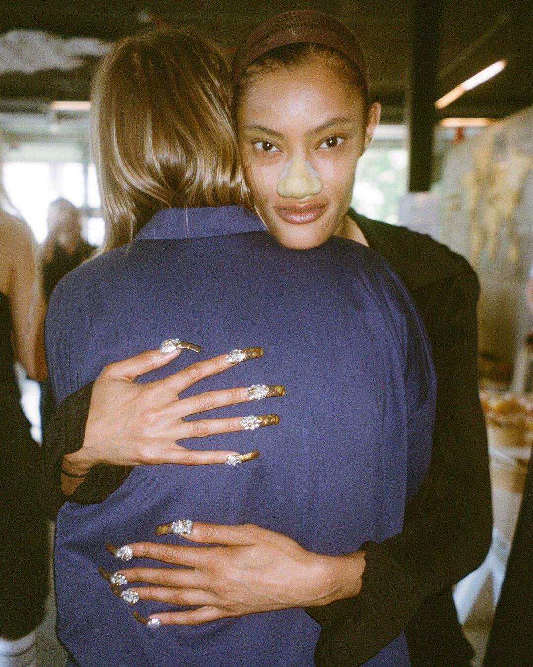 Two women embracing warmly in London, featuring detailed embellished long fingernails and a playful nose prosthetic.