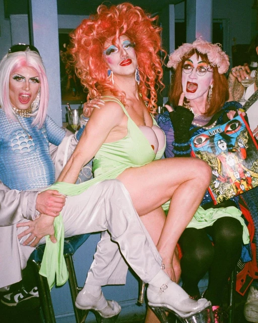 Three drag queens pose together indoors, wearing colorful outfits and wigs. The central figure sits on a stool in a vibrant green dress.