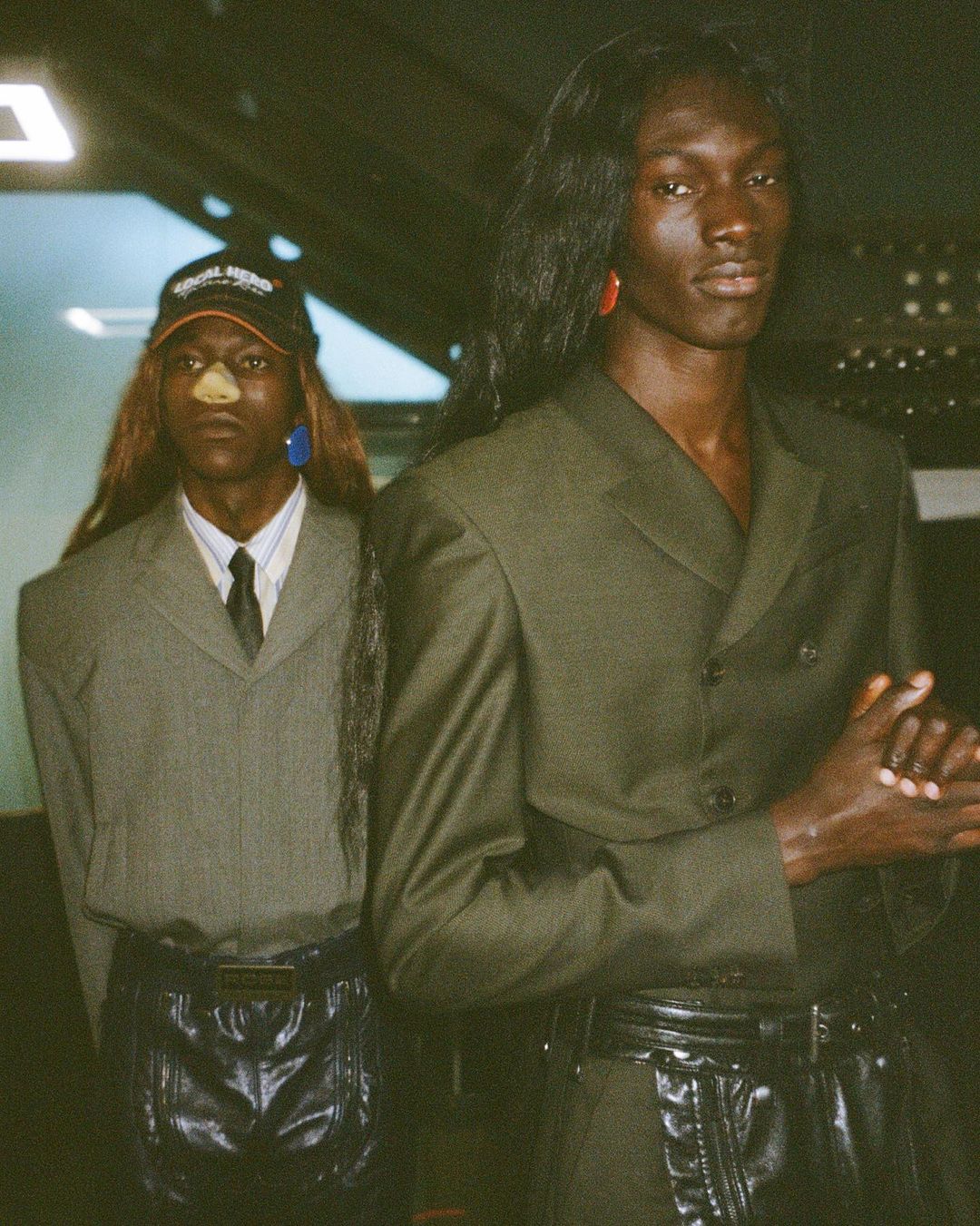 Two adult male fashion models pose in studio wearing matching green suits, black belts, and wigs against a dark background.