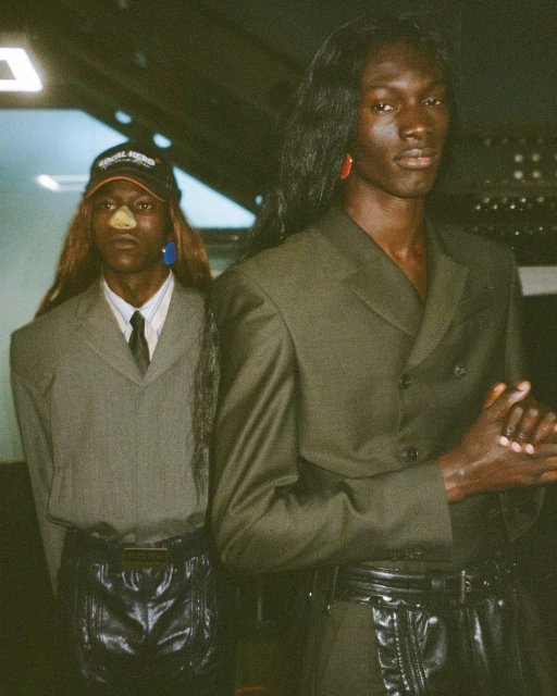 Two adult male fashion models pose in studio wearing matching green suits, black belts, and wigs against a dark background.