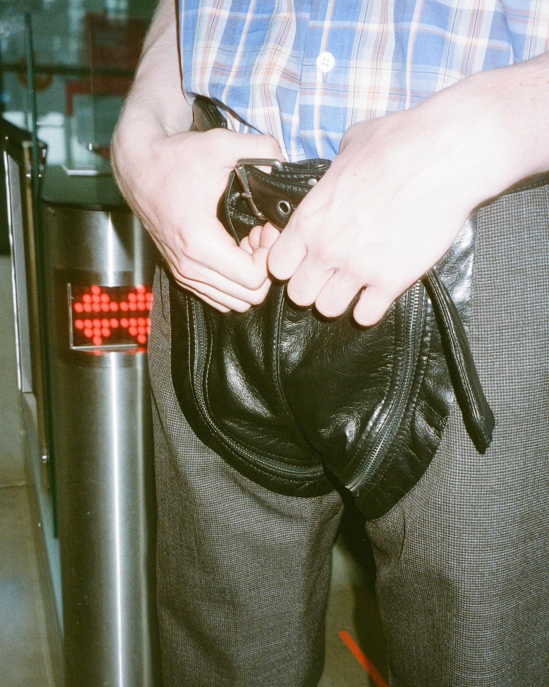 Adult male adjusts a black leather sporran-style bag worn over grey trousers, standing near a metal turnstile in an indoor setting.
