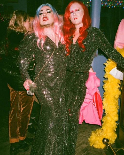 Two people in glittering gowns pose at an indoor event with bright indirect lighting; one has blue and pink hair, the other has red hair.