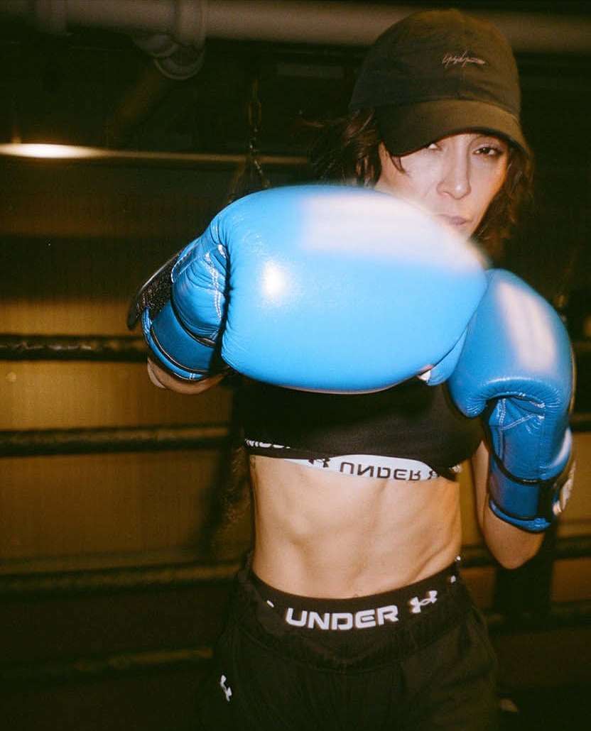 Woman in sportswear with boxing gloves in a boxing ring. She wears a sports bra, shorts, and a cap, posing in a low-lit indoor environment.
