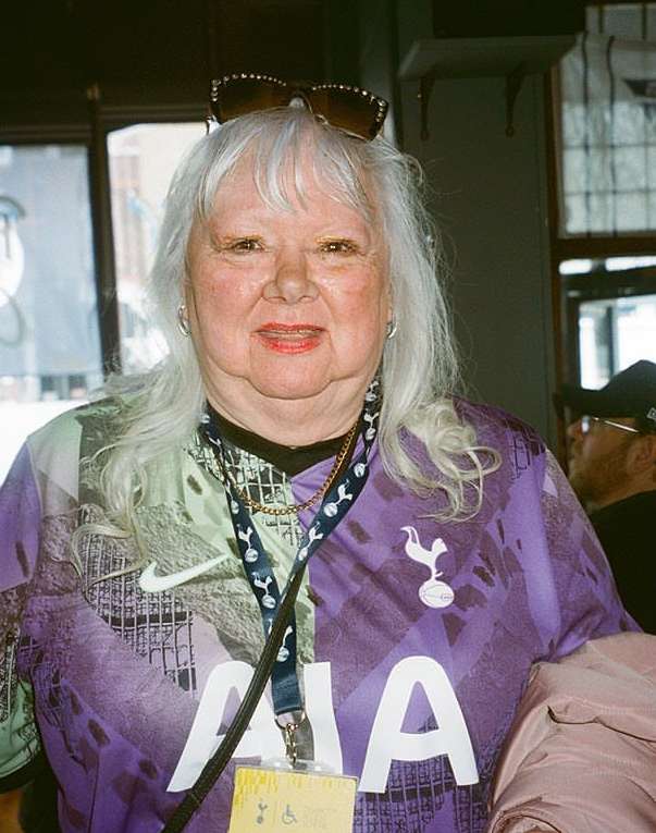 Elderly woman in soccer jersey with sunglasses on her head posing indoors; window and person visible in background.
