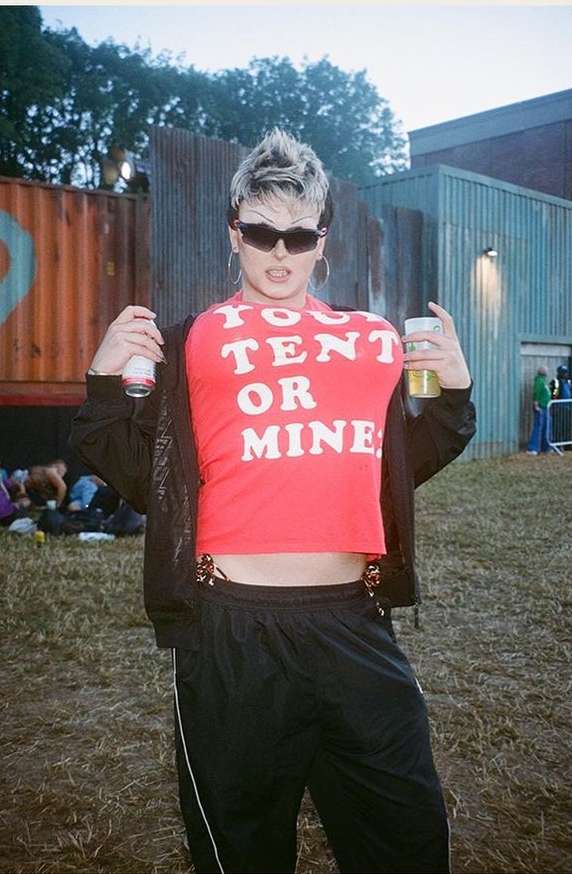 Person in a red printed t-shirt and black pants holds drinks outdoors. Casual style at an outdoor festival or event setting.