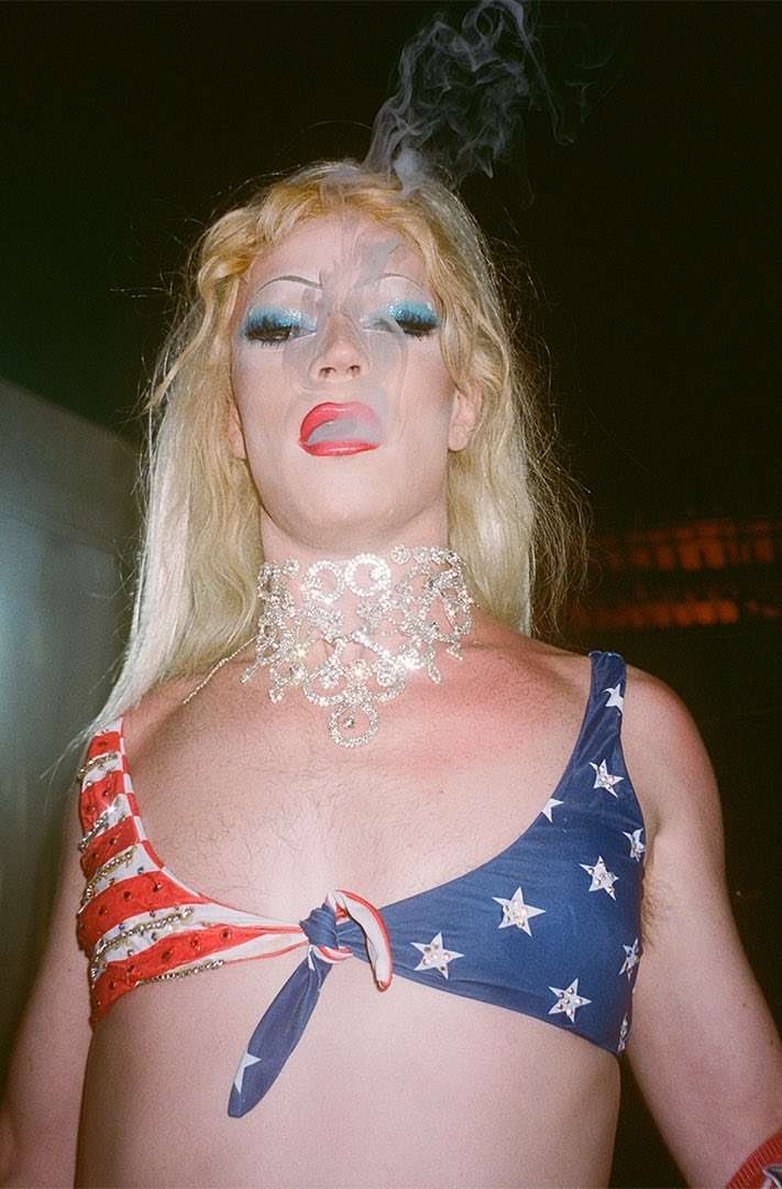 Close-up of a person with blonde hair, heavy makeup, and an American flag-themed bikini top smoking with a crystal choker.