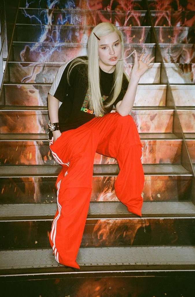 Young adult woman with blonde hair posing in black shirt and bright red Adidas track pants sitting on industrial stairs with a colorful graffiti-style mural.
