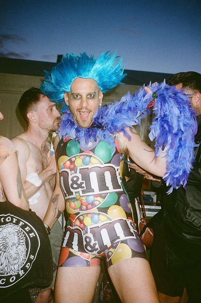 A person in a candy-themed outfit poses with a blue wig and feather boa outdoors, surrounded by a small group of people.