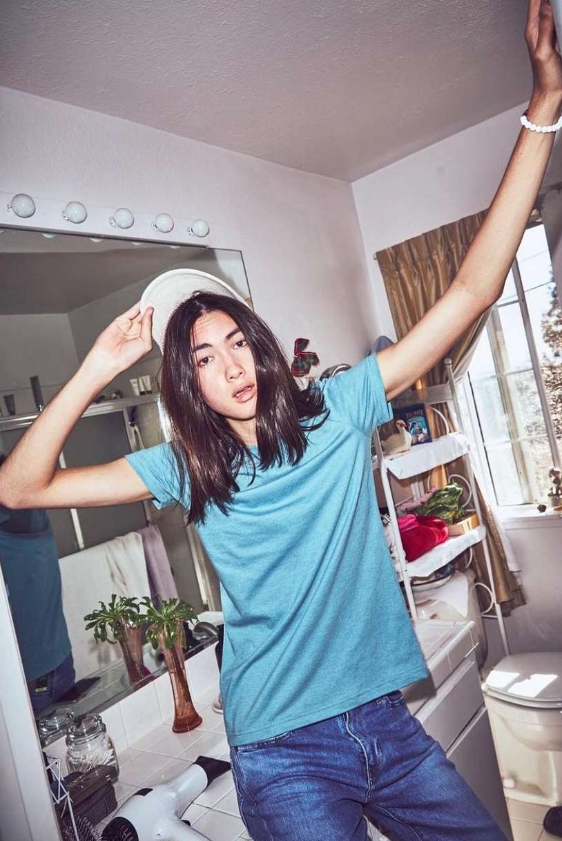 A young adult with long hair poses in a bathroom wearing a blue t-shirt and jeans, arms raised, lit by natural light.